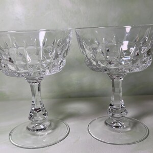 May include: Two clear glass coupe glasses featuring a faceted diamond-cut pattern on the bowl. Each glass has a short, sturdy stem and a wide, circular base. The design includes a textured geometric surface that reflects light across the clear glass.
