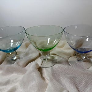 May include: Three glass coupes with wide, shallow bowls and clear stems. The left glass is light blue, the center glass is light green, and the right glass is a darker blue. Each piece features a smooth, rounded base and a simple, elegant design.