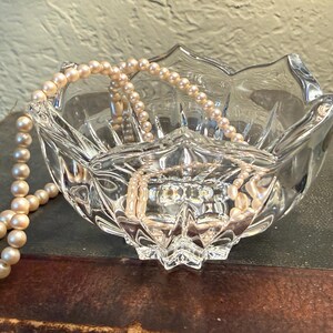 May include: A clear, faceted glass bowl with a scalloped edge, holding a strand of cream-colored pearls. The bowl sits on a dark surface, reflecting light and the pearls.