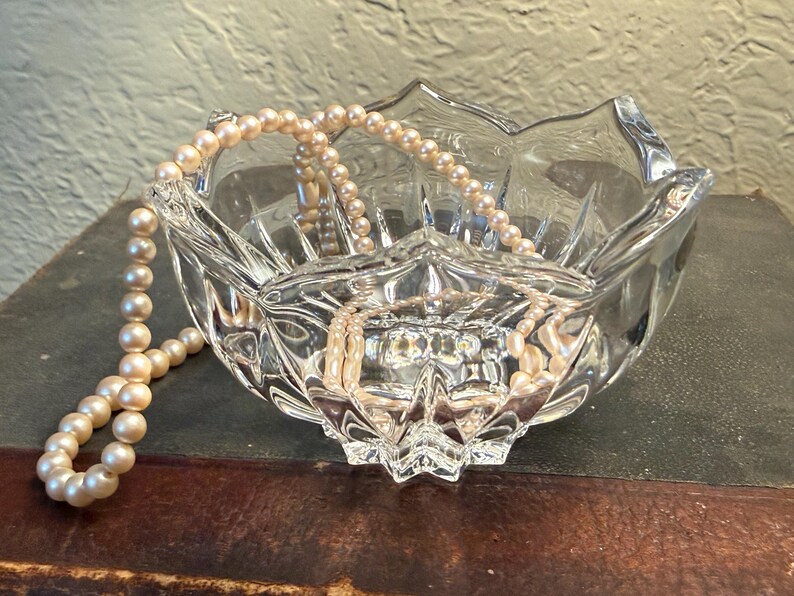 May include: A clear, faceted glass bowl with a scalloped edge, holding a strand of cream-colored pearls. The bowl sits on a dark surface, reflecting light and the pearls.