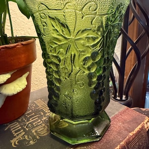 Puede incluir: Jarrón de vidrio verde con superficie texturizada y diseño de vid. El jarrón tiene una base hexagonal y una parte superior acampanada. El jarrón está sobre un libro antiguo.