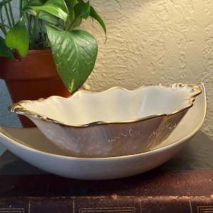 May include: A cream-coloured serving set comprising a scalloped-edge bowl with gold trim, nested on a crescent-shaped dish. The set is displayed on a dark surface, with a potted plant in the background.