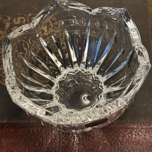May include: A clear, faceted crystal bowl with a flower-like design. The bowl has a scalloped rim and a starburst pattern on the interior base. The bowl is sitting on a dark brown surface.
