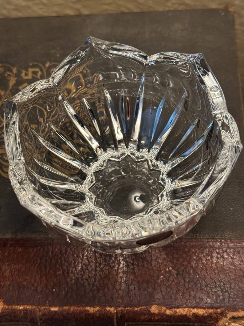 May include: A clear, faceted crystal bowl with a flower-like design. The bowl has a scalloped rim and a starburst pattern on the interior base. The bowl is sitting on a dark brown surface.