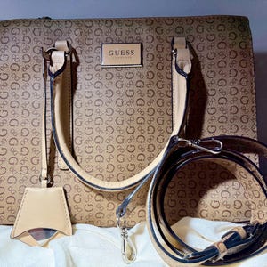May include: A tan Guess handbag with a repeating 'G' pattern. The bag has beige handles, a detachable shoulder strap, and a small tag. The Guess logo is displayed on a rectangular plaque on the front of the bag.