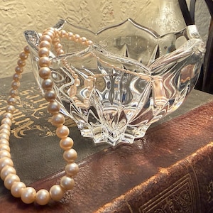 May include: A clear, cut-glass bowl with a scalloped edge, resting on a vintage book. A strand of peach-colored pearls drapes over the bowl. The scene is lit with warm, natural light, creating a classic, elegant aesthetic.