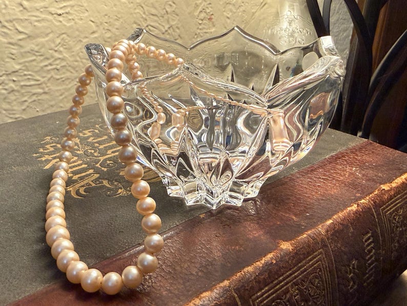May include: A clear, cut-glass bowl with a scalloped edge, resting on a vintage book. A strand of peach-colored pearls drapes over the bowl. The scene is lit with warm, natural light, creating a classic, elegant aesthetic.