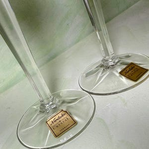 May include: Two clear crystal wine glasses with tall, slender stems and circular bases. Each base features a gold-colored square sticker with the text Noritake Crystal Germany printed in black, indicating the brand and origin of the glassware.