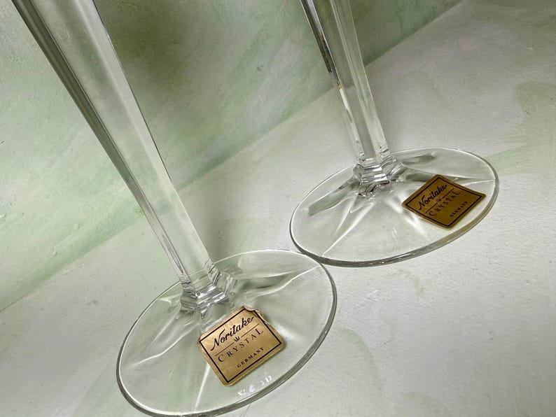 May include: Two clear crystal wine glasses with tall, slender stems and circular bases. Each base features a gold-colored square sticker with the text Noritake Crystal Germany printed in black, indicating the brand and origin of the glassware.