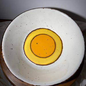 May include: A white ceramic bowl with a speckled surface and a brown rim. The center features a bright yellow circle with a brown border, creating a striking contrast. The bowl is likely used for serving food.