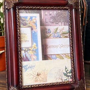 May include: A rectangular, dark red picture frame with ornate gold-colored detailing. The frame holds three vintage birthday greeting cards with floral and architectural imagery. The words "Happy Birthday" and "Birthday Greetings" are visible on the cards.