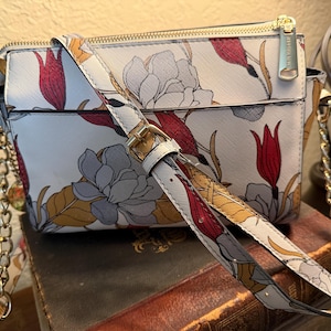 May include: A white crossbody bag with a floral pattern featuring red, yellow, and gray flowers. The bag has a gold zipper and chain strap, and a matching patterned strap. The bag is sitting on top of a stack of books.