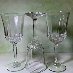 May include: Three clear crystal wine glasses with a faceted stem and a starburst pattern at the base of the bowl. Two glasses stand upright on circular bases, while the center glass is inverted, resting on its rim with the base facing upward.