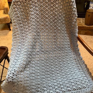 Puede incluir: Una manta de ganchillo azul claro sobre una silla de madera. La manta presenta un patrón de puntada de concha detallado y un borde festoneado. La manta hecha a mano es un artículo de decoración suave y acogedor.