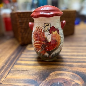 Puede incluir: Un recipiente de cerámica con tapa y asas rojas, decorado con un motivo de gallo. El gallo está representado en tonos rojos, naranjas y marrones. El fondo es blanquecino con escritura. El recipiente está sobre una superficie de madera.