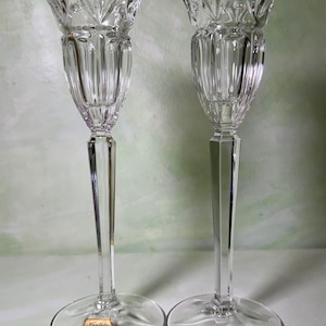 May include: A pair of clear crystal candle holders featuring a tall, slender stem and a flared, faceted top with vertical ribbing. Each base displays a gold-colored sticker with the text Noritake Crystal Germany printed in black lettering.