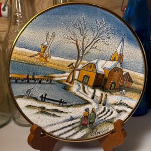 May include: A round decorative plate with a winter scene featuring a church, windmill, and figures walking on a snowy path. The plate has a gold rim and is displayed on a wooden stand. The artwork is signed "V. Uziaro".