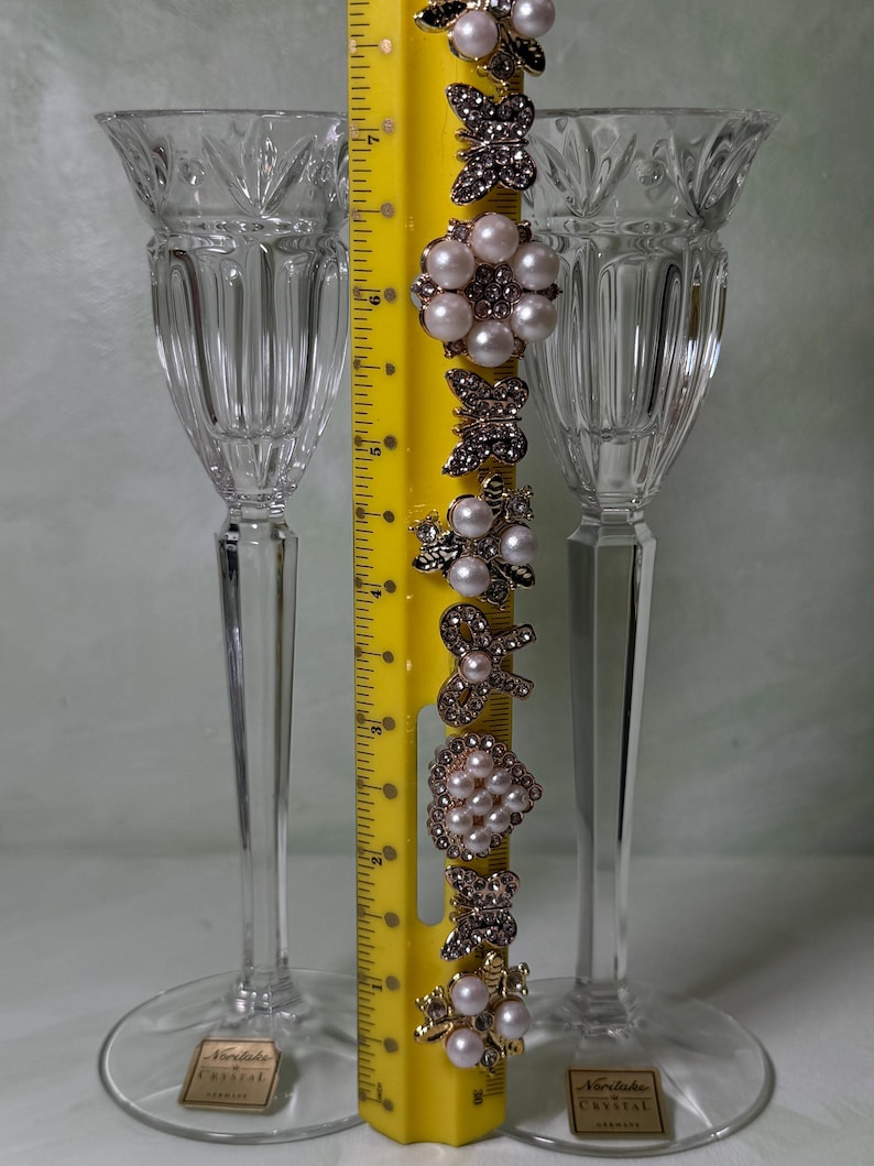 May include: Two clear crystal stemmed glasses with vertical fluting and a yellow ruler measuring approximately 7 inches in height. A vertical row of decorative metal embellishments featuring faux pearls and rhinestones in butterfly and floral shapes.