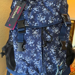 May include: A navy blue backpack with a floral pattern of small white and light blue flowers. The backpack has black buckles, a drawstring closure, and a side pocket. The tag reads "super backpack".