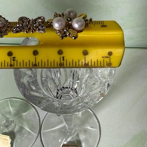 May include: Two clear crystal glasses with a faceted bowl and a stem, each featuring a Noritake Crystal label. A yellow ruler is placed across the top, showing the width of the decorative metal embellishments with pearls and rhinestones.