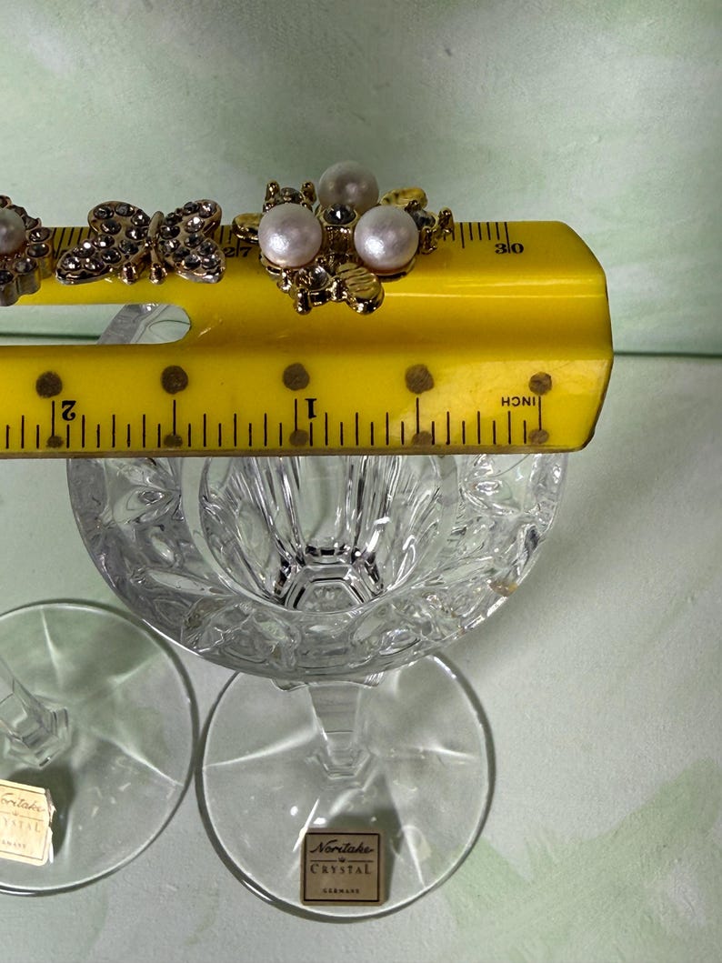 May include: Two clear crystal glasses with a faceted bowl and a stem, each featuring a Noritake Crystal label. A yellow ruler is placed across the top, showing the width of the decorative metal embellishments with pearls and rhinestones.