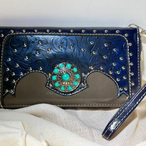 May include: A blue leather wallet with a decorative turquoise stone centerpiece. The wallet features embossed floral patterns, silver studs, and a wrist strap. The bottom section is a muted gray color.