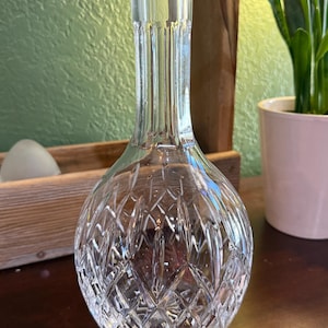 May include: A clear glass vase with a round base and a long, narrow neck. The vase has a diamond-cut pattern on the lower half and vertical lines on the neck. The vase is sitting on a dark brown surface.