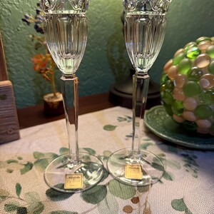 May include: A pair of clear crystal candle holders with a fluted bowl design and a faceted stem. Each piece features a circular base and a gold-colored sticker with the text Noritake Crystal printed on the front of the base.