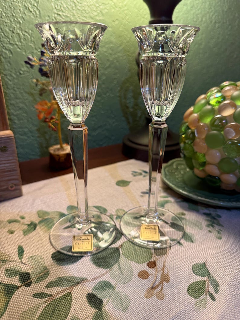 May include: A pair of clear crystal candle holders with a fluted bowl design and a faceted stem. Each piece features a circular base and a gold-colored sticker with the text Noritake Crystal printed on the front of the base.