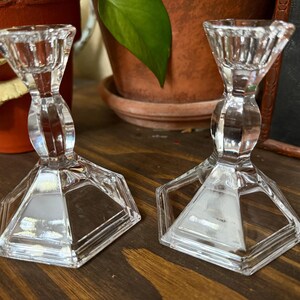 May include: Two clear glass candle holders with a faceted design. The candle holders have a hexagonal base and a flared top. They are displayed on a wooden surface.