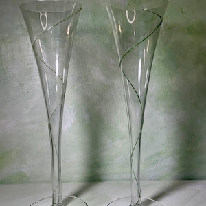 May include: A pair of tall, clear glass champagne flutes featuring a delicate, thin green glass thread spiraling around the exterior of each bowl and stem. The glasses have a wide, circular base and a slender, elongated conical shape.
