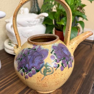 May include: A handcrafted ceramic teapot with a yellow base and a decorative handle. The teapot features purple floral designs and a brown rim and spout. The handle is adorned with a textured design. A unique piece for tea lovers.