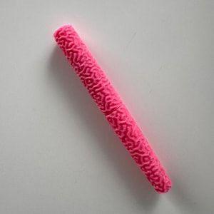 Puede incluir: Un objeto cilíndrico de color rosa brillante con un patrón texturizado similar a un cerebro. El objeto está acostado en diagonal sobre una superficie blanca. El color es un rosa vibrante y sólido.