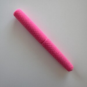 Puede incluir: Un objeto cilíndrico de color rosa brillante con un patrón tejido texturizado. El objeto está colocado en diagonal sobre una superficie blanca. Parece ser un estuche o soporte para un objeto pequeño.