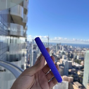 Puede incluir: Un objeto cilíndrico texturizado, de un tono azul vibrante, se sostiene contra un telón de fondo de un paisaje urbano bajo un cielo azul claro. La superficie del objeto presenta un patrón repetitivo. La estética general sugiere un tema urbano moderno.