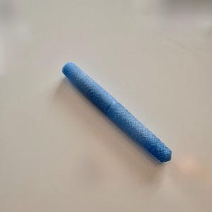 Puede incluir: Un objeto cilíndrico azul claro con un patrón tejido texturizado. El objeto está acostado en diagonal sobre una superficie blanca. El objeto parece ser un bolígrafo o un instrumento de escritura similar.