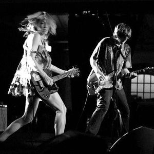 May include: Black and white image of two people playing electric guitars on a stage. The person on the left is wearing a patterned dress and the person on the right is wearing a shirt and jeans. The stage is dark with a window in the background.