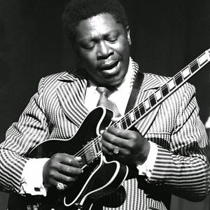 Pode incluir: Fotografia a preto e branco de um músico a tocar uma guitarra elétrica preta. O músico está a usar um fato às riscas, camisa branca com colarinho e gravata. A imagem é um plano médio, com foco no músico e na guitarra.