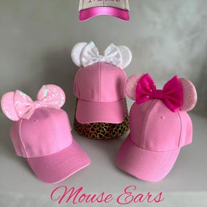 May include: Pink baseball caps with attached mouse ears and bows. The caps have a curved peak and are displayed with a matching cap featuring the word "Mouse". The text "Mouse Ears Base Ball Cap" is also visible.