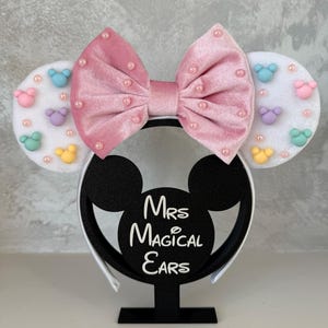 May include: A pink velvet bow headband with pearl accents and white ears decorated with pastel-colored mouse shapes. The headband is displayed on a black stand with the text "Mrs Magical Ears".