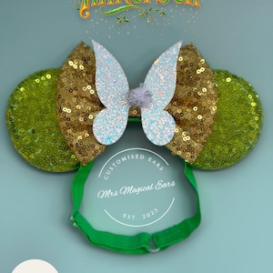 May include: A green headband with gold sequin ears and a gold sequin bow with a glittery butterfly in the centre. The text "Tinkerbell" is at the top. The text "Mrs Magical Ears" is on the headband.