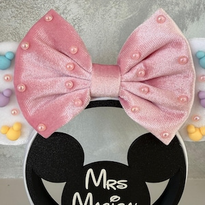 May include: A pink velvet bow headband with pearl accents. The headband features black Mickey Mouse ears with the text "Mrs Magical Ears" in white. The ears are attached to a white headband.