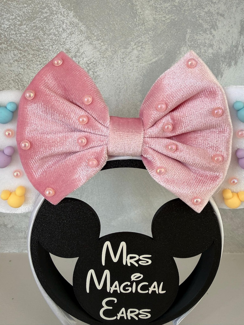 May include: A pink velvet bow headband with pearl accents. The headband features black Mickey Mouse ears with the text "Mrs Magical Ears" in white. The ears are attached to a white headband.