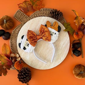 May include: A white headband with a sequined orange bow and character ears. One ear features a black and white cartoon face. The other ear has an orange bow. The headband is decorated with autumn-themed elements.
