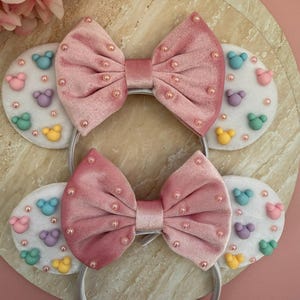 May include: Two headbands with pink velvet bows and white ear-shaped accents. The accents feature colorful mouse-shaped designs and small pink beads. The headbands are white and sit on a light-colored surface.