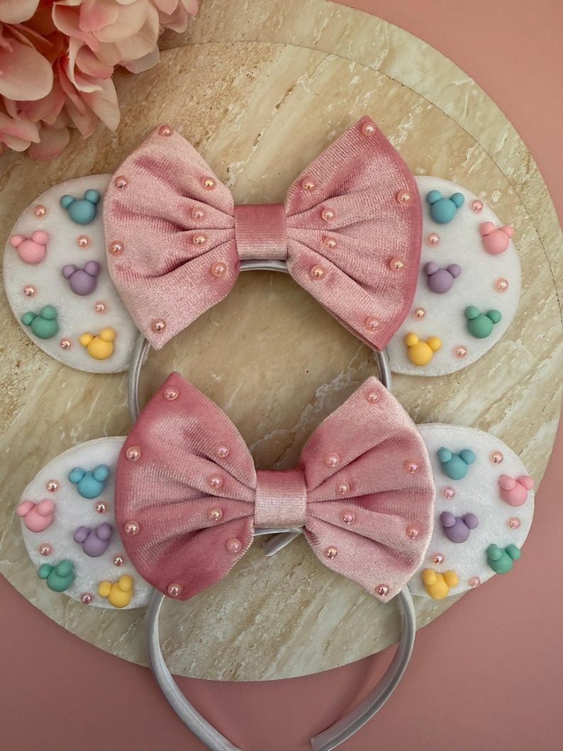 May include: Two headbands with pink velvet bows and white ear-shaped accents. The accents feature colorful mouse-shaped designs and small pink beads. The headbands are white and sit on a light-colored surface.