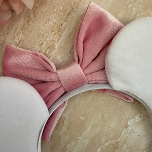 May include: A pink velvet bow headband with white plush ears. The headband features a large, soft pink bow in the center and two round, white plush ears on either side. The headband is set against a marble background.
