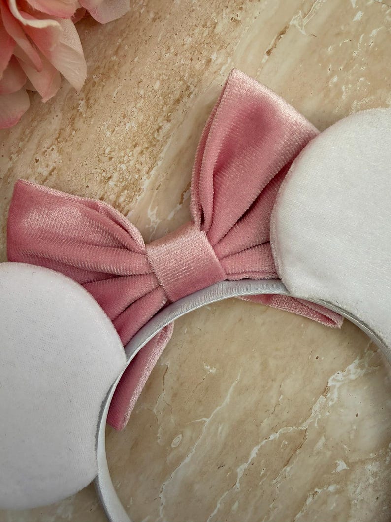 May include: A pink velvet bow headband with white plush ears. The headband features a large, soft pink bow in the center and two round, white plush ears on either side. The headband is set against a marble background.