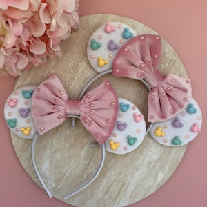 May include: Pink velvet bow headbands with pearl accents and white felt ears decorated with colorful cartoon character shapes. The headbands are on a round, marble-like surface against a pink background.