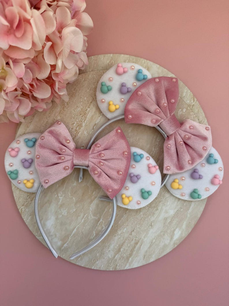 May include: Pink velvet bow headbands with pearl accents and white felt ears decorated with colorful cartoon character shapes. The headbands are on a round, marble-like surface against a pink background.
